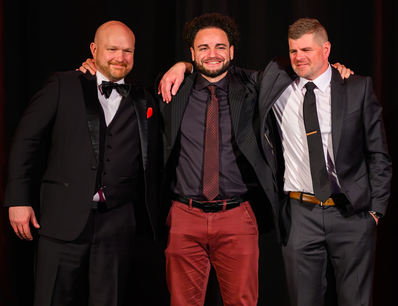 Three men standing on stage with their arms around each others' shoulders.